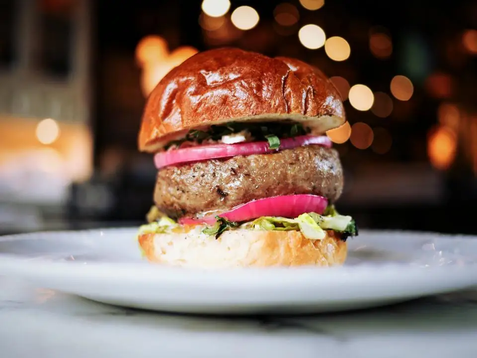 A burger with pickled red onion on a white plate, warm lights behind